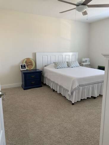 La imagen muestra un dormitorio sereno con una cama blanca, paredes blancas y moqueta beige, una mesilla de noche, un ventilador de techo y un cartel "Be my guest" en la pared.